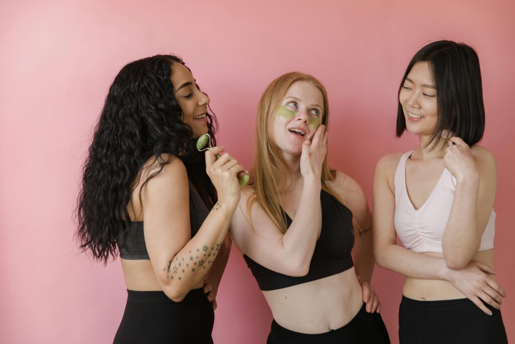 Three women in gym clothing enjoying a skincare routine with face rollers in a studio with a pink background.